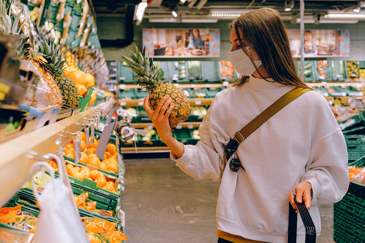 Woman in mask chooses pineapple in a supermarket for safe shopping during pandemic.