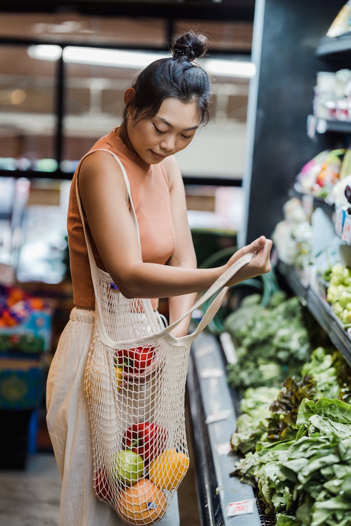 Asian woman in a supermarket selecting fruits and vegetables using a reusable mesh bag.